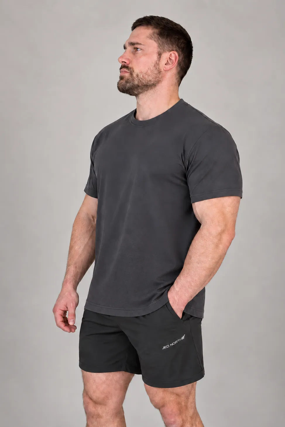 A man wearing a gray athletic training tee and black shorts by Jed North standing against a neutral background with a confident posture