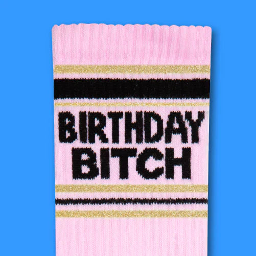 Pink knit birthday bitch crew socks with black and gold stripes featuring bold black lettering on the front