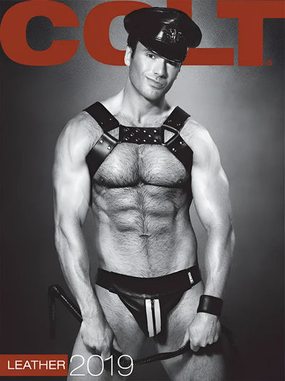 Muscular man in a leather cap and harness stands against gray backdrop.