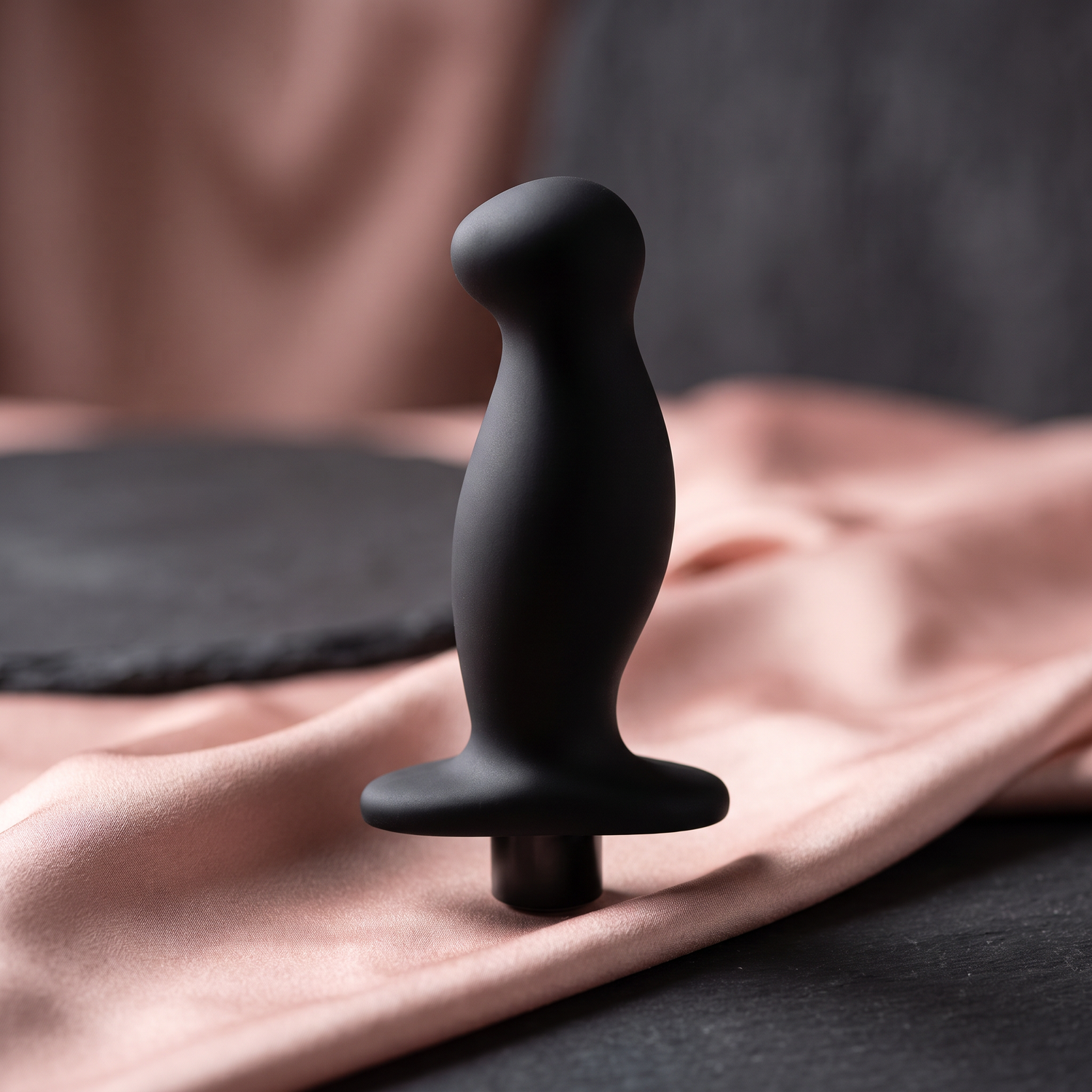 Black silicone wellness device on a blush and charcoal studio background with open space for cropping