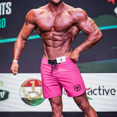 A bodybuilder in bright pink shorts poses on stage with a competition number.