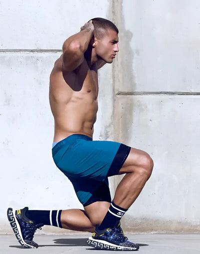 Shirtless man kneeling in teal athletic shorts before a concrete wall background.