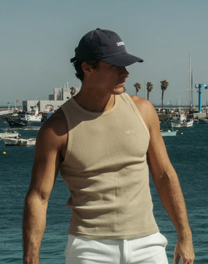 Beige ribbed tank top with small chest logo, shown with white pants and a marina backdrop.