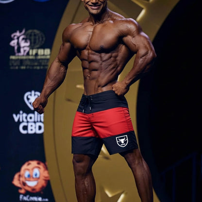 A muscular man wearing red and black MetroMaleWear physique board shorts with a white bull logo on the left leg