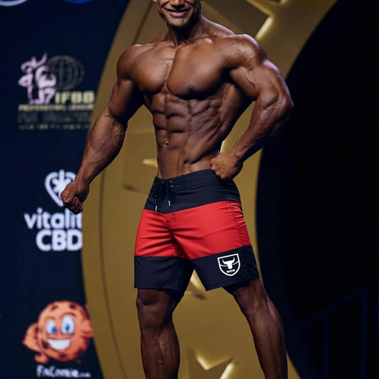 A muscular man wearing red and black MetroMaleWear physique board shorts with a white bull logo on the left leg