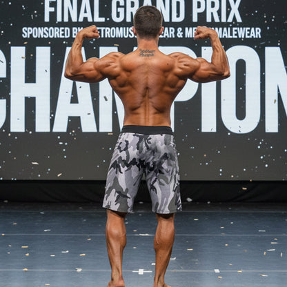 A muscular man wearing metroMalewear physique board short camo 4706-60 camouflage pattern with black waistband stands on stage