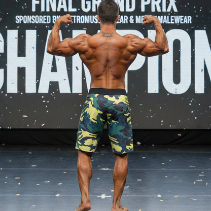 A muscular man stands on stage wearing MetroMaleWear physique board shorts in yellow green camo pattern
