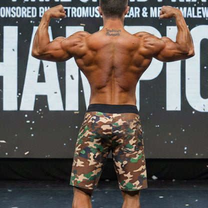 A muscular man wearing metroMalewear physique board shorts in green camouflage pattern with brown and beige tones
