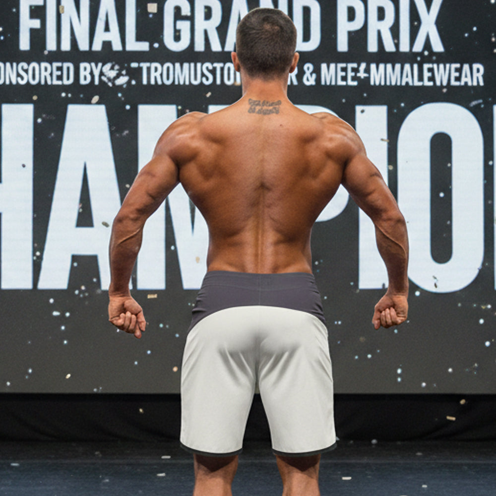 A muscular man stands on stage wearing gray MetroMaleWear MetroMuscleWear Cordoba physique board shorts with a dark gray waistband