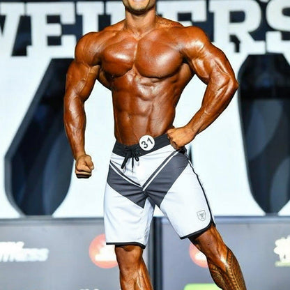 A muscular man wearing white MetroMaleWear MetroMuscleWear Mark Physique board shorts with logo on lower right leg