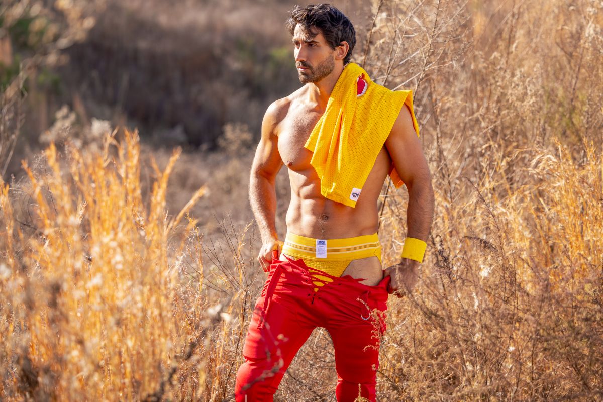 A man wearing bright red athletic pants with yellow trim and a yellow BIKE athletic jockstrap visible from the back side