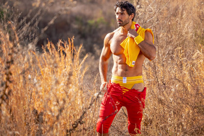 A man wearing a yellow bike athletic jockstrap with torn red shorts and a bright yellow shirt in a sports setting