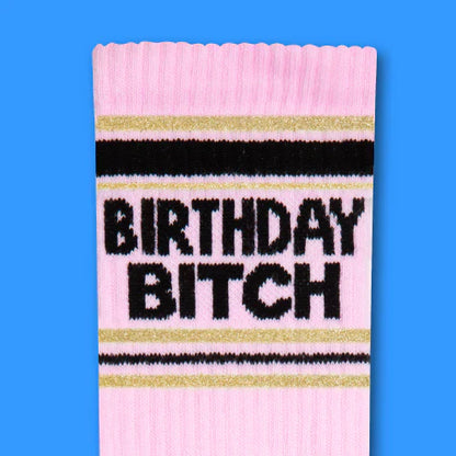 Pink knit birthday bitch crew socks with black and gold stripes featuring bold black lettering on the front