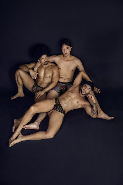 Three men pose in matching black chili pepper print CandyMan 99875 thongs with a dark patterned design against black background