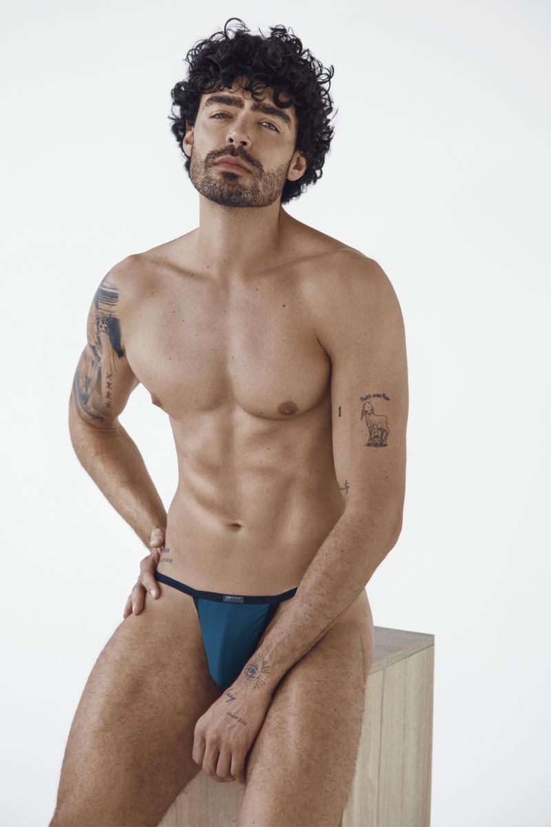 A muscular man with dark curly hair and tattoos wearing petrol blue briefs while sitting on a block.
