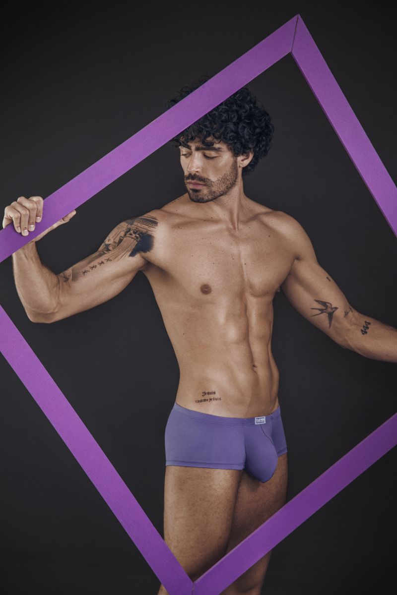 A man with curly hair wearing lilac trunks, posing inside a purple rectangular frame.