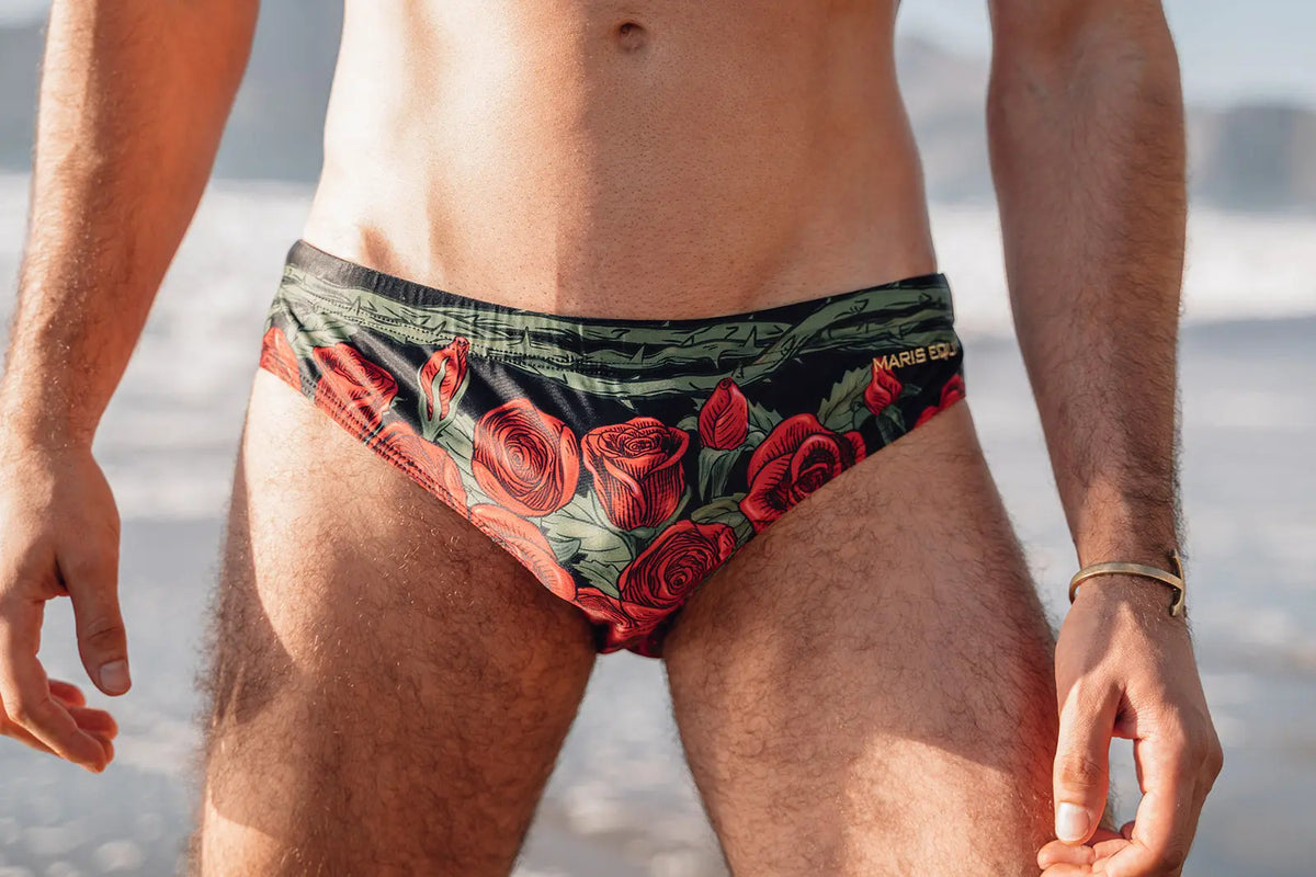 Men's swim briefs in black with red rose print featuring a low-rise cut and Maris Equi branding worn by a model at the beach.