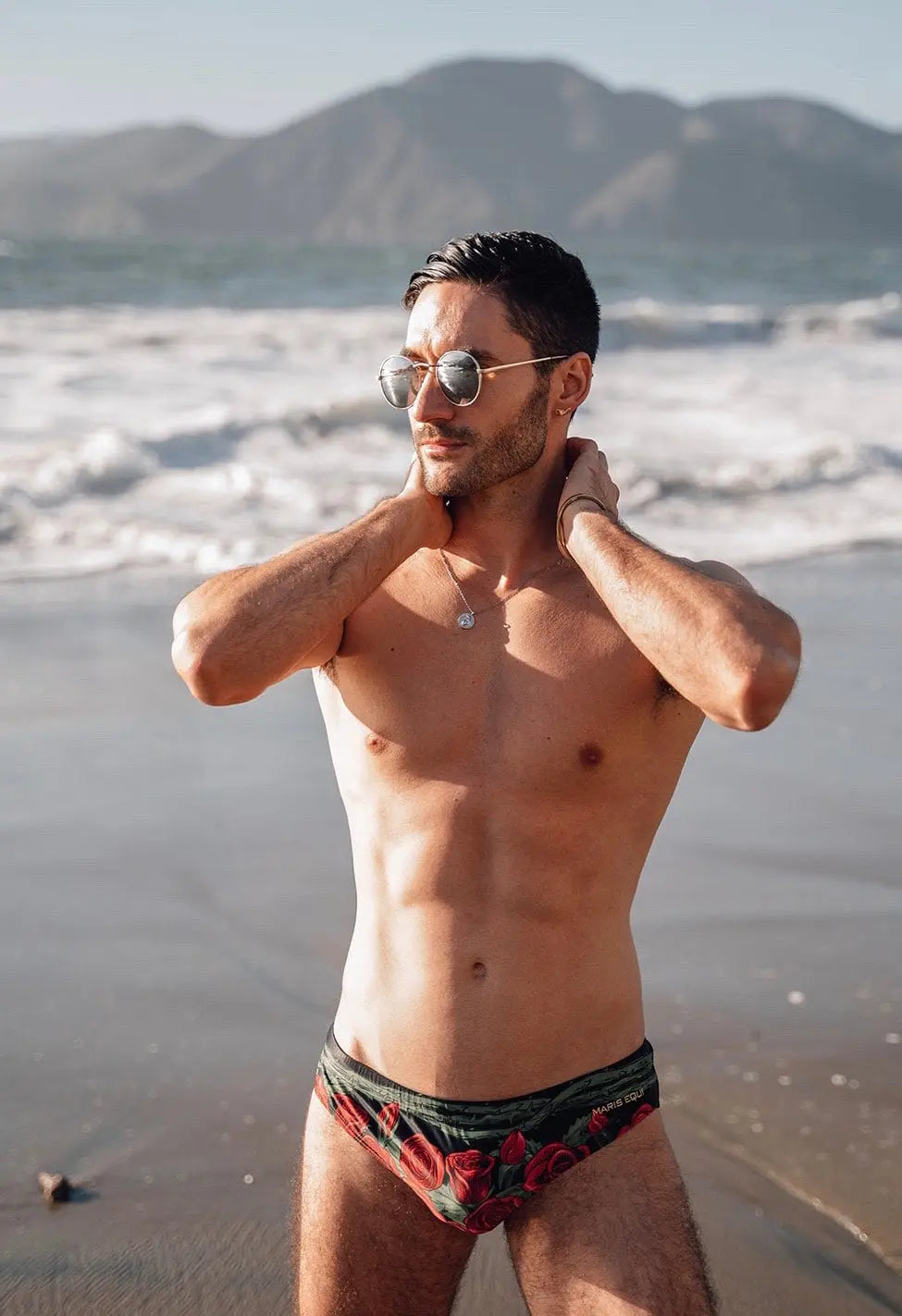 Men's swim briefs in black with red rose print made by Maris Equi shown on a model at the beach.