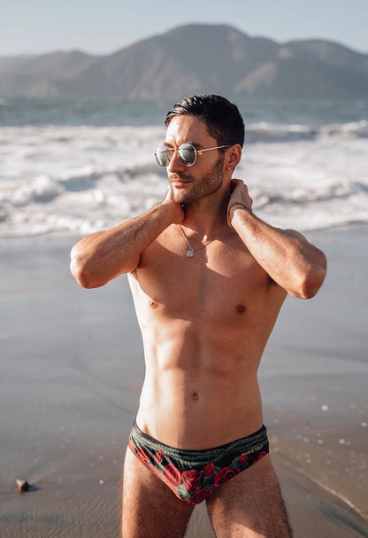 Men's swim briefs in black with red rose print made by Maris Equi shown on a model at the beach.