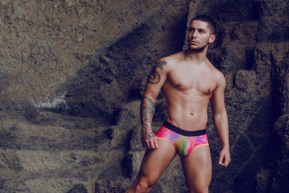 The man is wearing mob aero brief swim trunks in vibrant rainbow colors with a black waistband