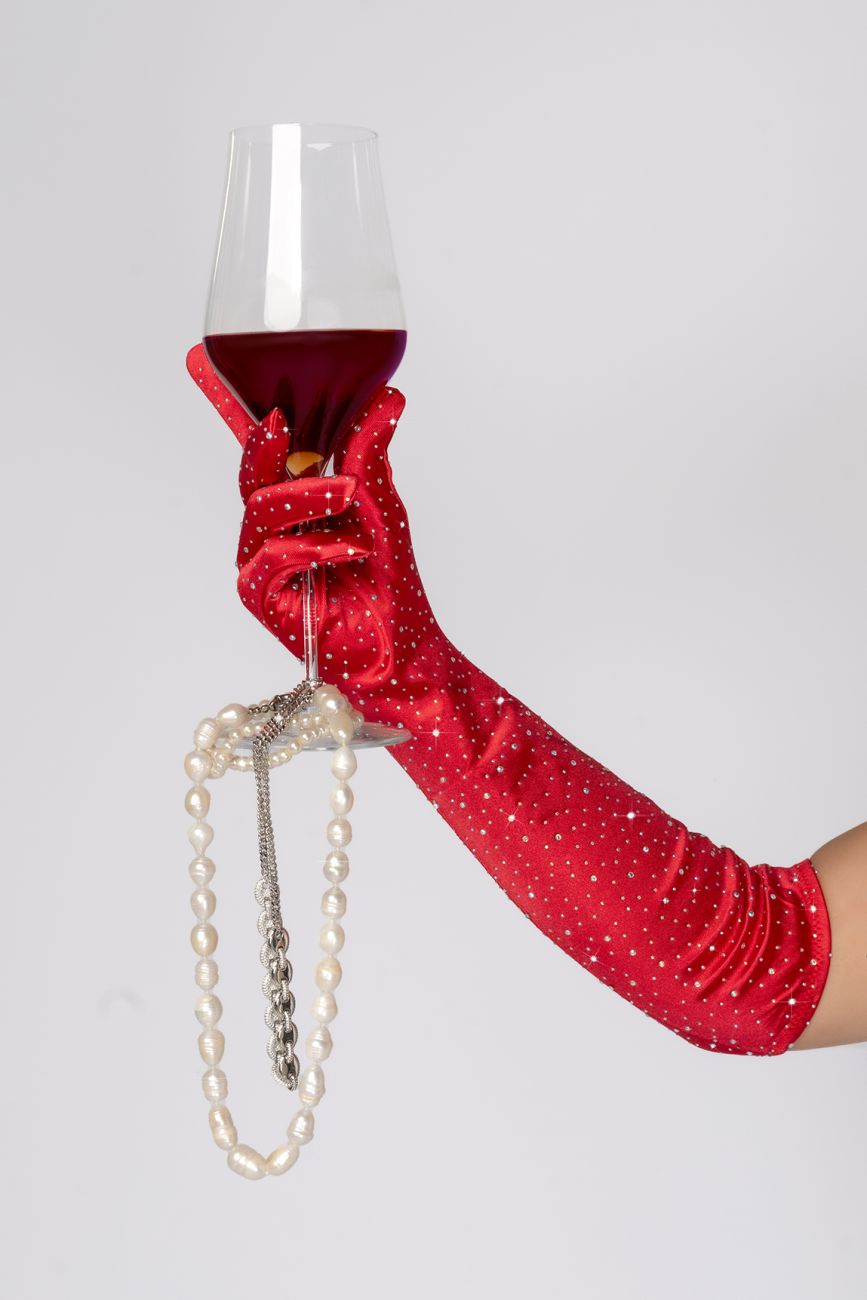 A hand wearing mapale 135 red satin rhinestone opera gloves holds a wine glass with a pearl necklace on the stem
