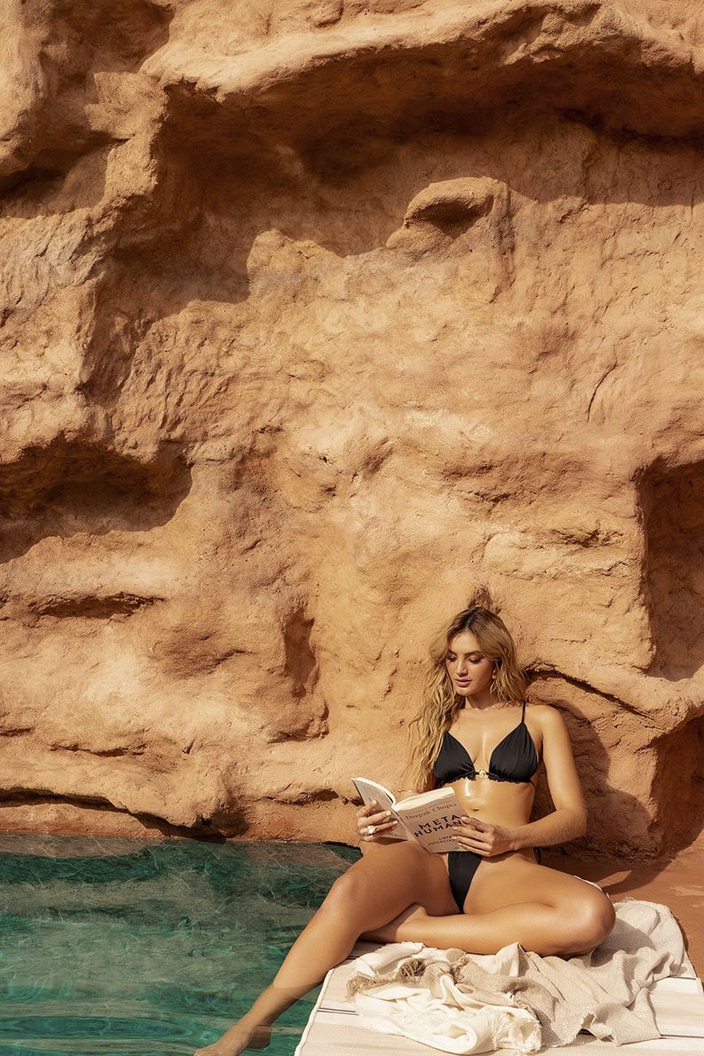A woman sits by the poolside wearing a black Mapale 67166 two piece swimsuit reading a book titled milia