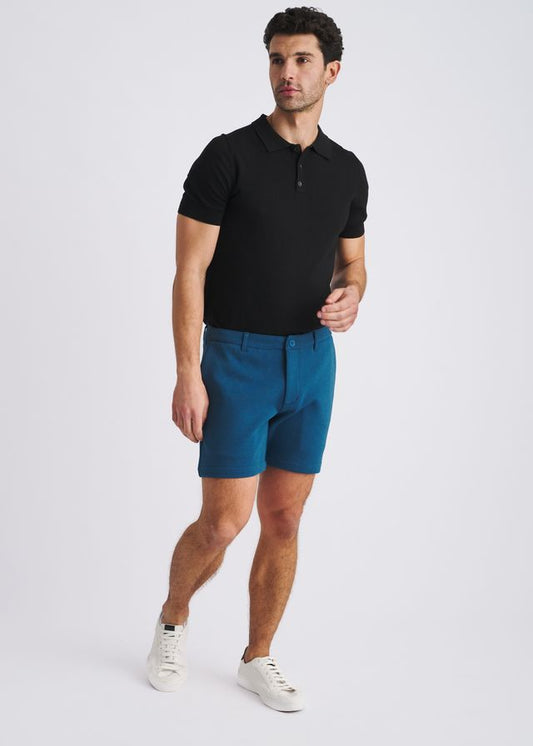 POSTMARC petrol stretch knit chino shorts worn with a black polo and white sneakers on a light grey background.