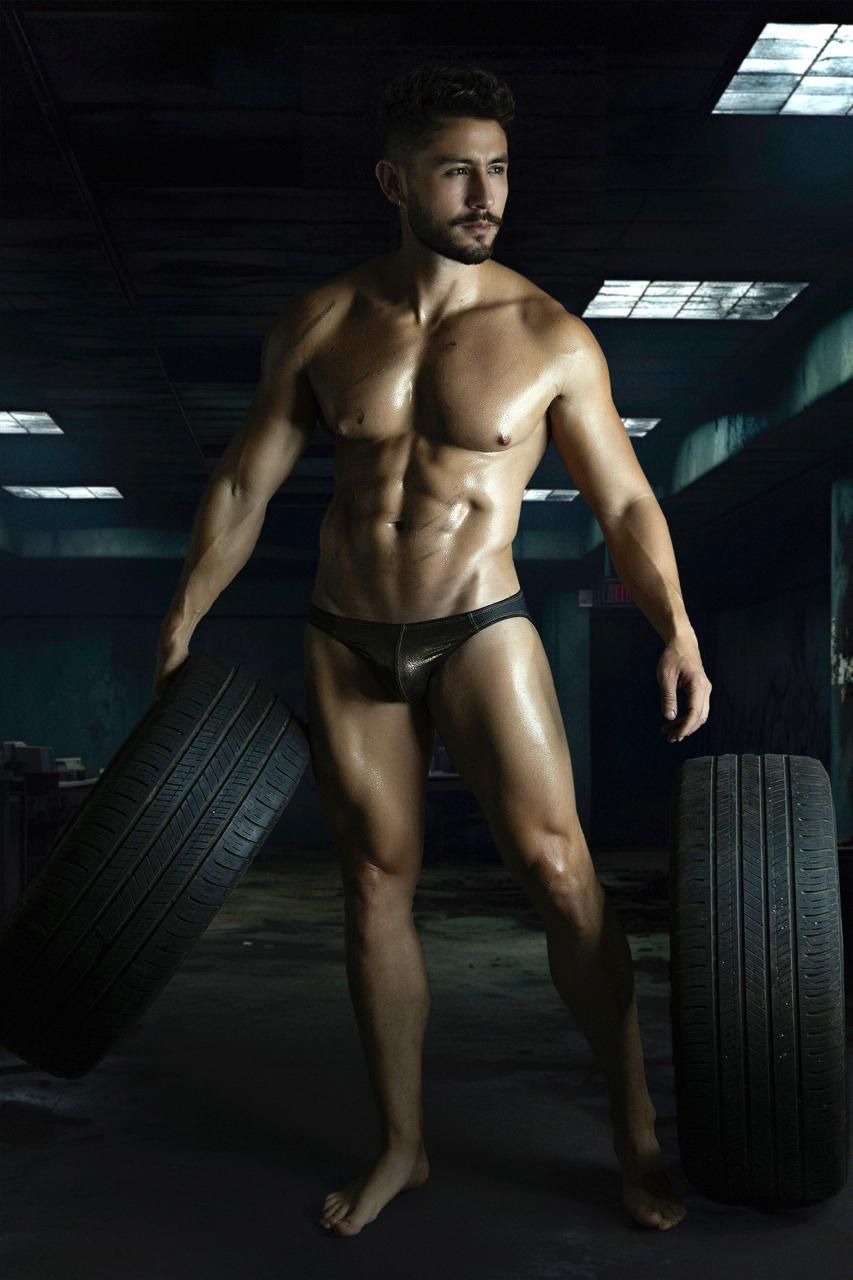 A muscular man wearing black shiny form-fitting Pikante PIK 2121 Gold Cup briefs stands barefoot between tires
