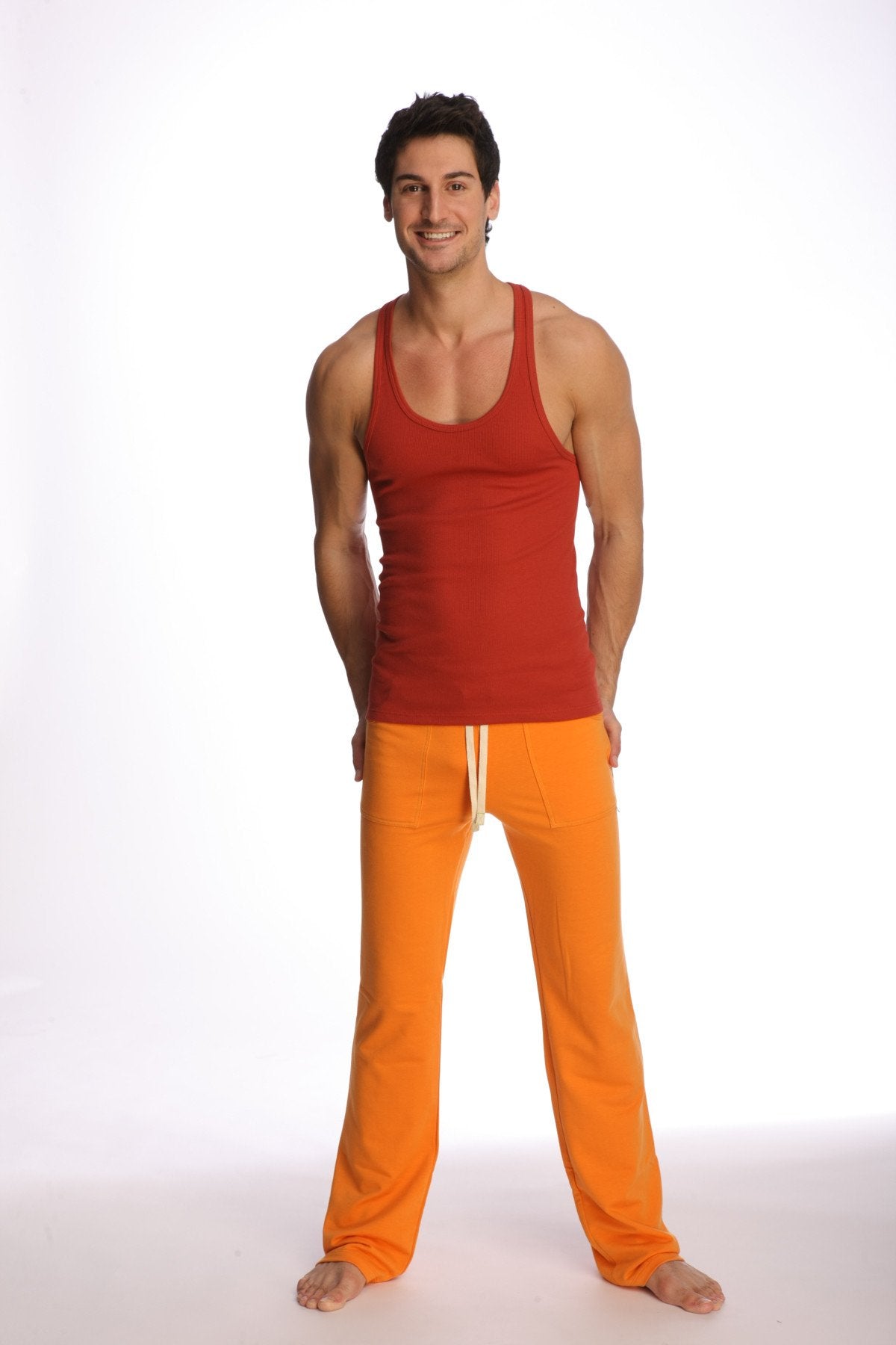A man wearing a cinnabar racer-back yoga tank in red with orange pants featuring white drawstrings