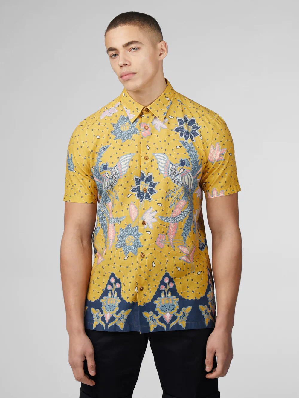 Yellow short-sleeved button-up shirt with intricate blue pink and white floral and bird patterns on a dotted background