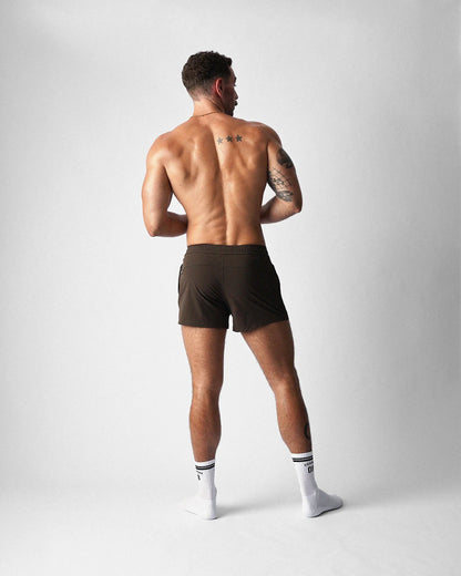 Rear view of a person wearing teamm8 Bolt 3" brown gym shorts and white crew socks, on a grey background.