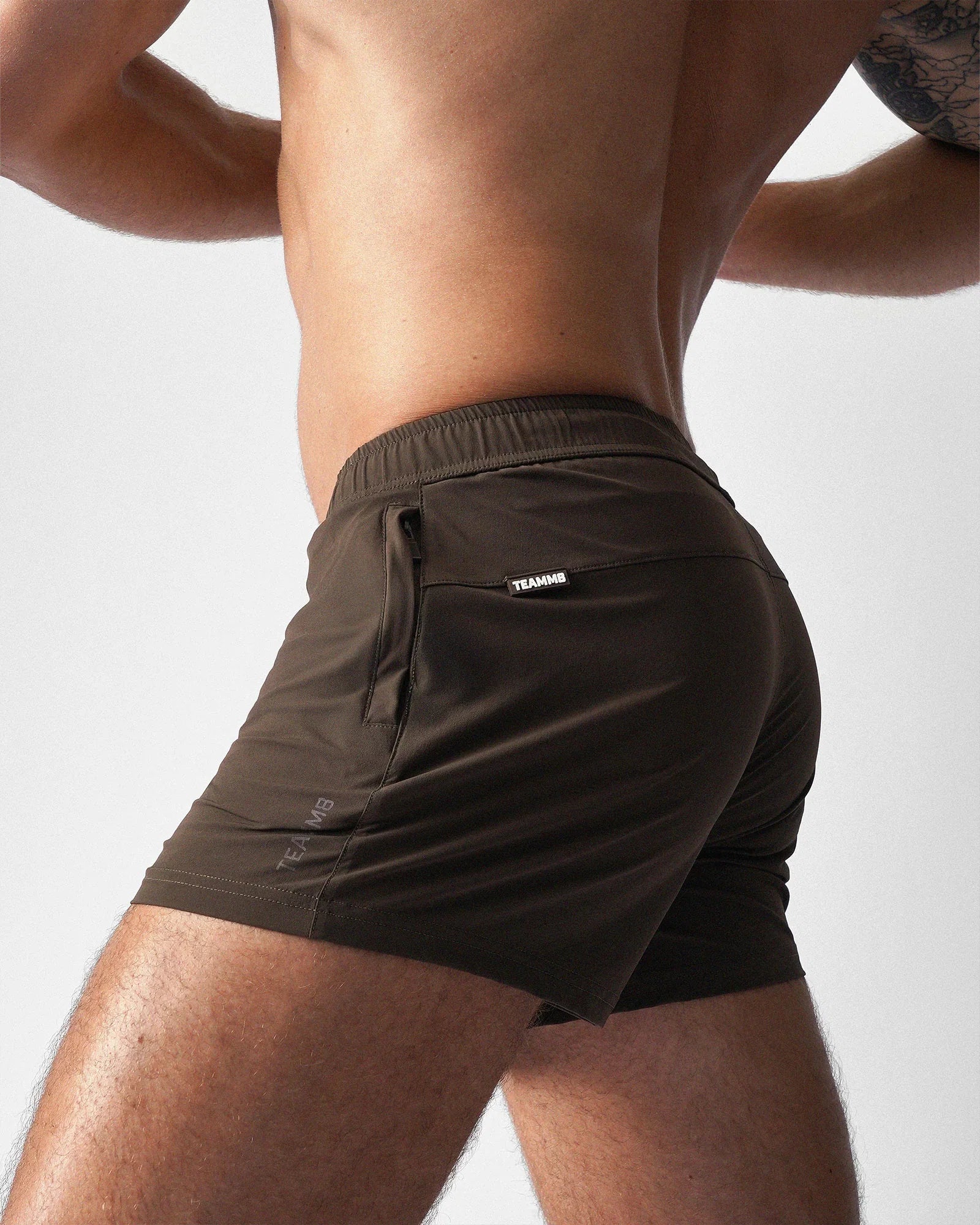 Side view of a person wearing dark brown teamm8 Bolt 3" gym shorts with a zippered pocket and a small white logo tag.