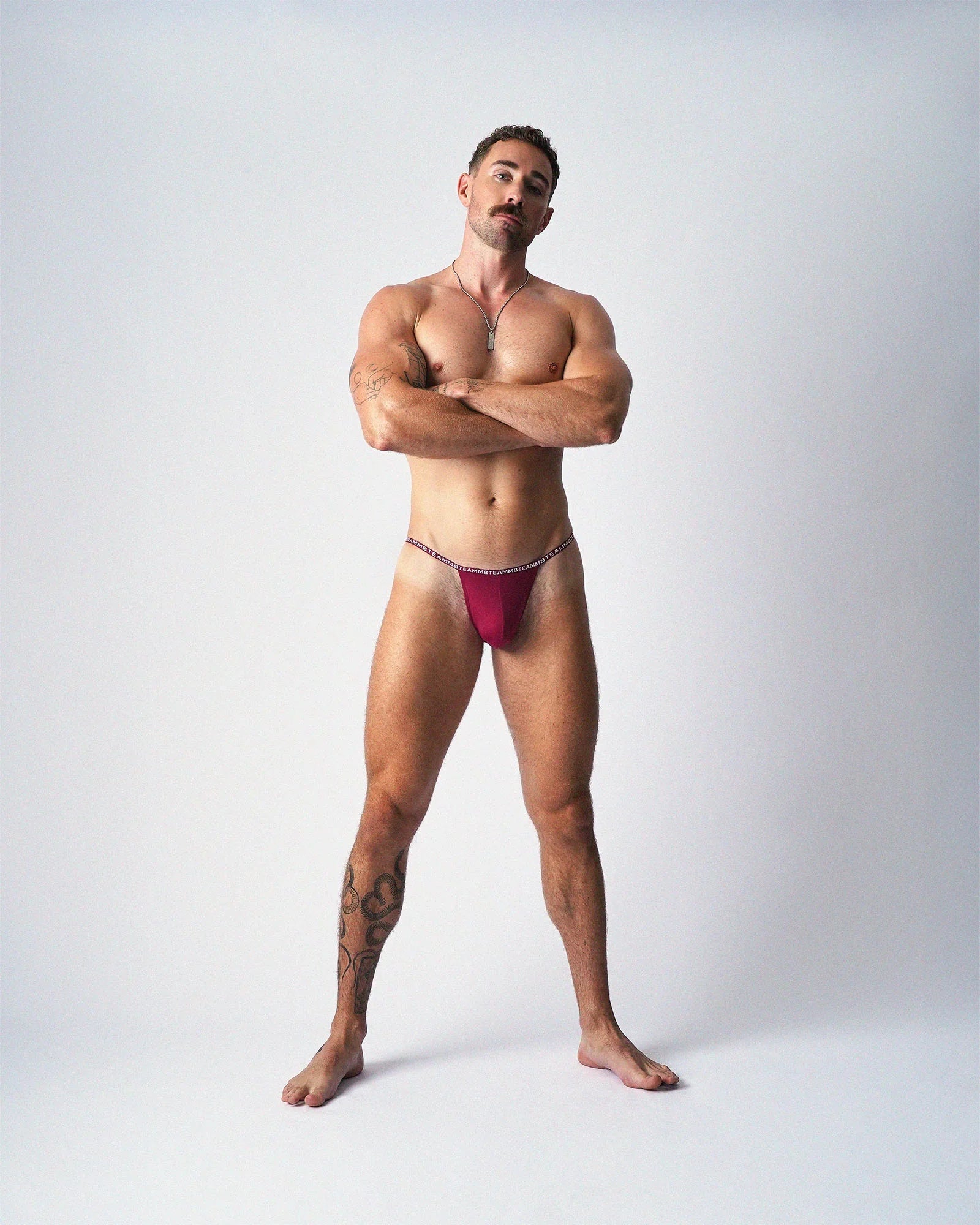 A standing man with arms crossed wears maroon teamm8 Glide Micro Briefs against a plain white background.