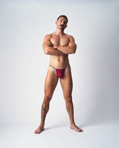 A standing man with arms crossed wears maroon teamm8 Glide Micro Briefs against a plain white background.