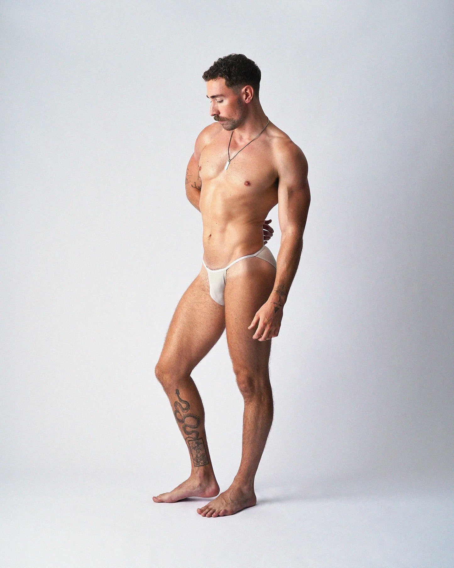 A standing person wears light beige micro briefs against a plain grey background, viewed in three-quarter profile.