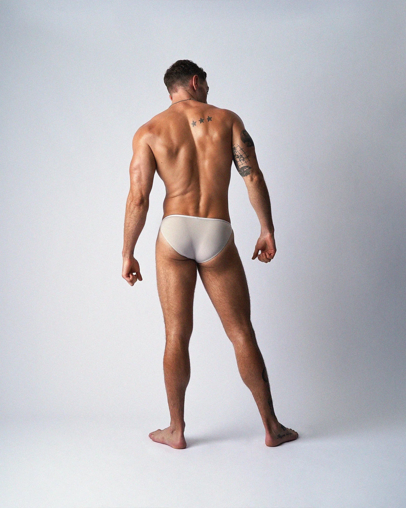 Rear view of a person wearing light beige micro briefs against a plain grey background.
