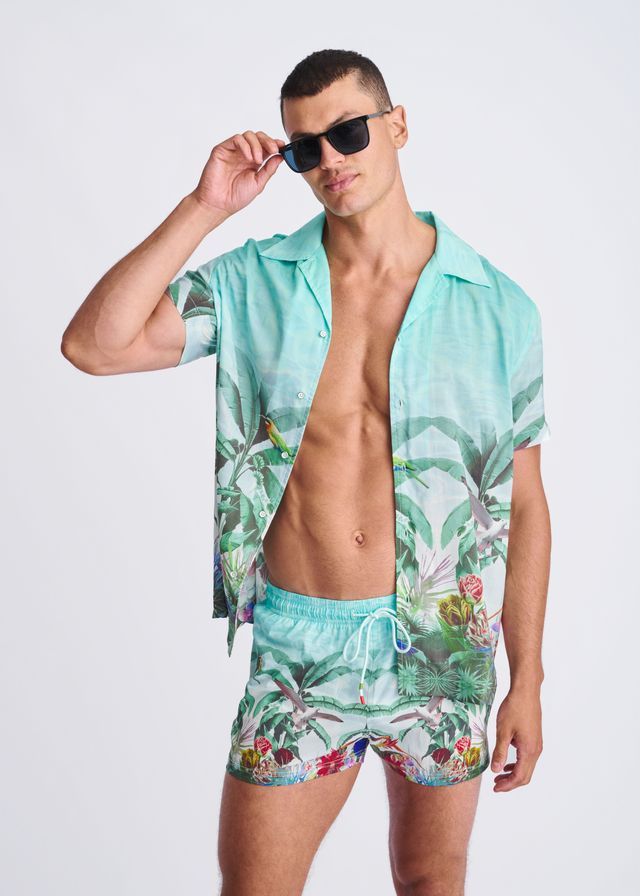 A vibrant turquoise tropics print swim shorts with mesh lining featuring palm leaves and birds paired with matching shirt