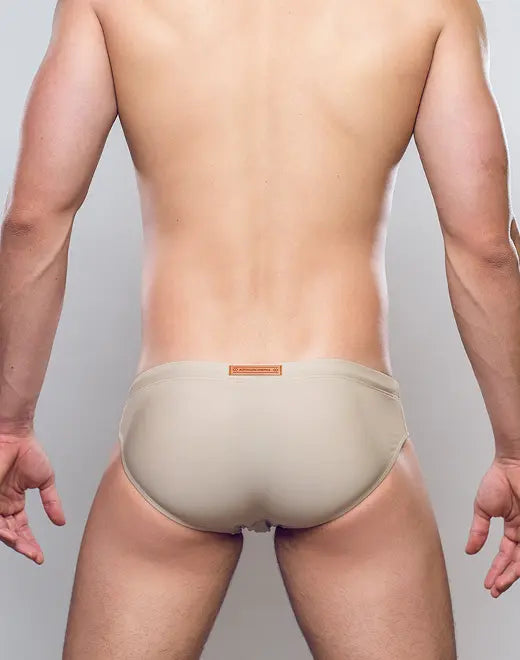 Beige form-fitting v10 core bikini swim trunks with a subtle orange brand tag at the waistband