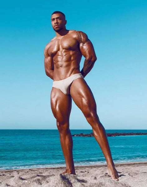 A muscular man stands on a sandy beach wearing white v10 core bikini briefs showcasing the front side