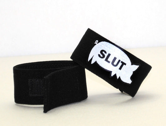 Two black fabric cock rings with hook-and-loop fasteners; one features a white pig silhouette with "SLUT" printed.