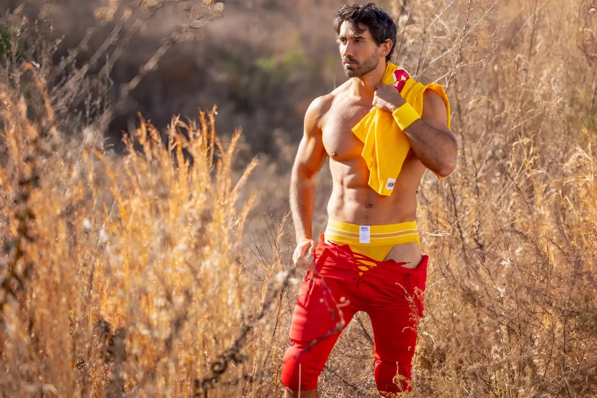 A man wearing a yellow bike athletic jockstrap with torn red shorts and a bright yellow shirt in a sports setting