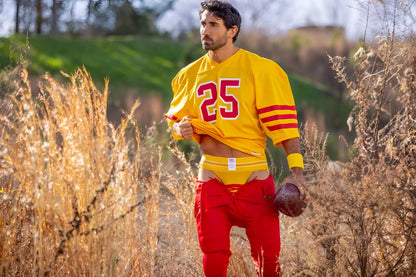 A man wearing a yellow jockstrap with a vibrant yellow and red football jersey and pants.