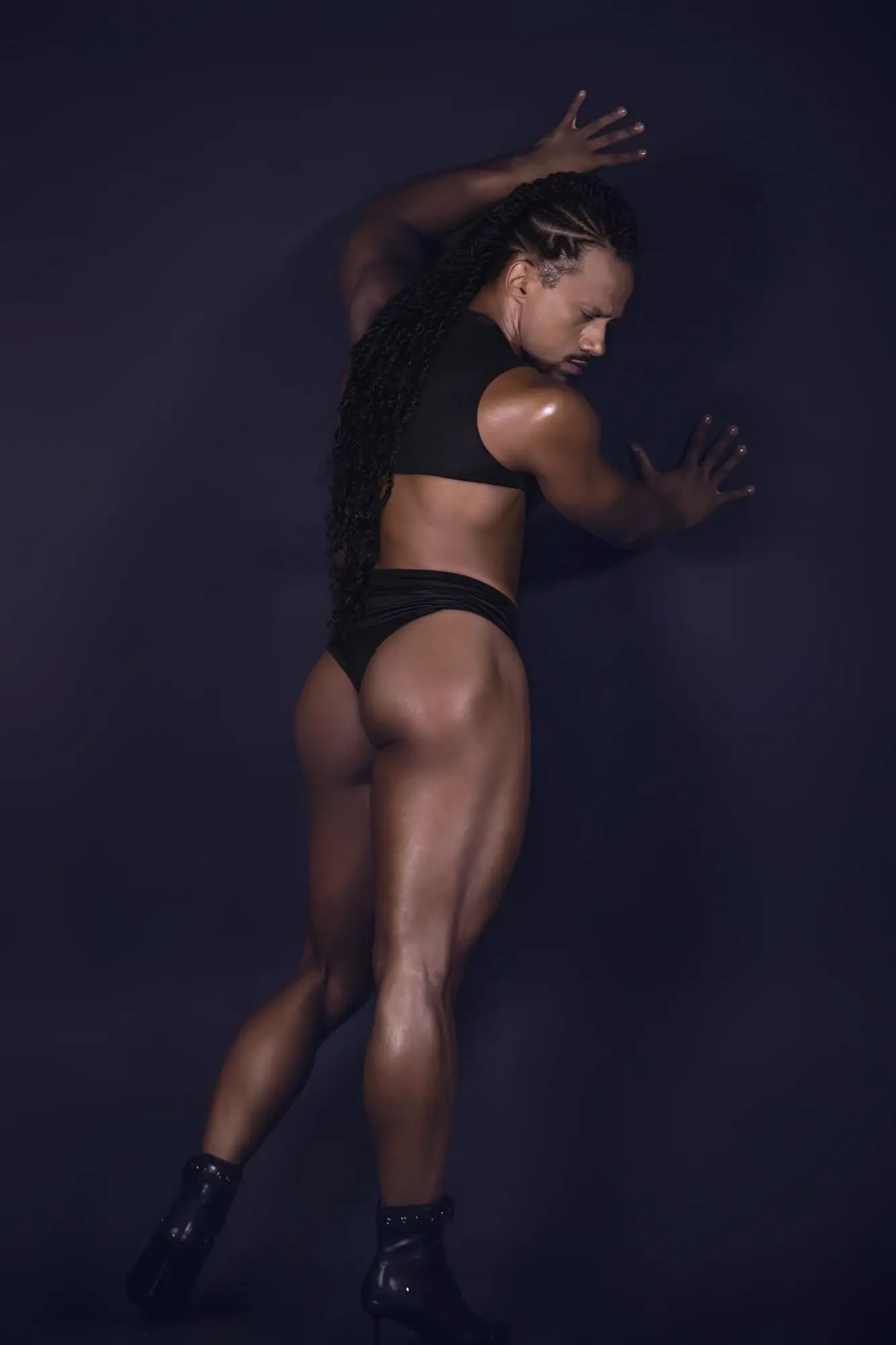 A muscular individual wearing a black candy man 99783 one shoulder bodysuit paired with glossy black heeled boots