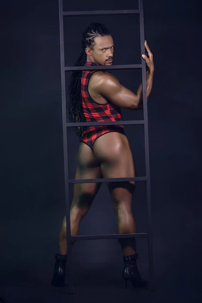 A muscular man with long braided hair wearing a red and black plaid sleeveless candyMan 99800 lumberjack bodysuit stands behind a dark metal ladder