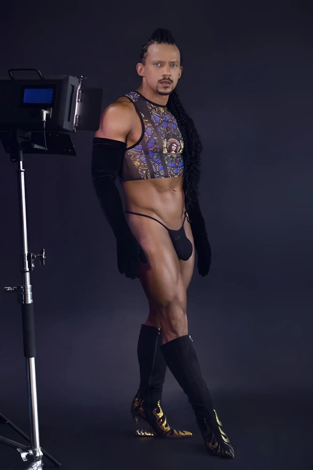 A man wearing a candyMan 99805 stained glass tank top in black with gold patterns and matching thigh-high boots