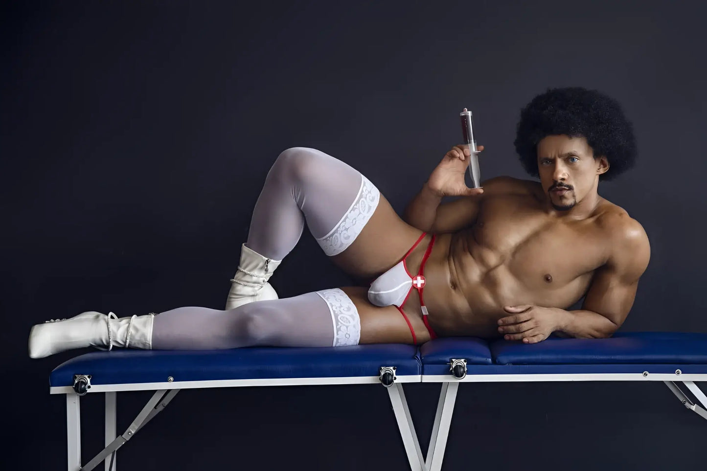 A muscular man wearing CandyMan 99833 murse thongs in red and white with white lace thigh-high stockings on a blue table