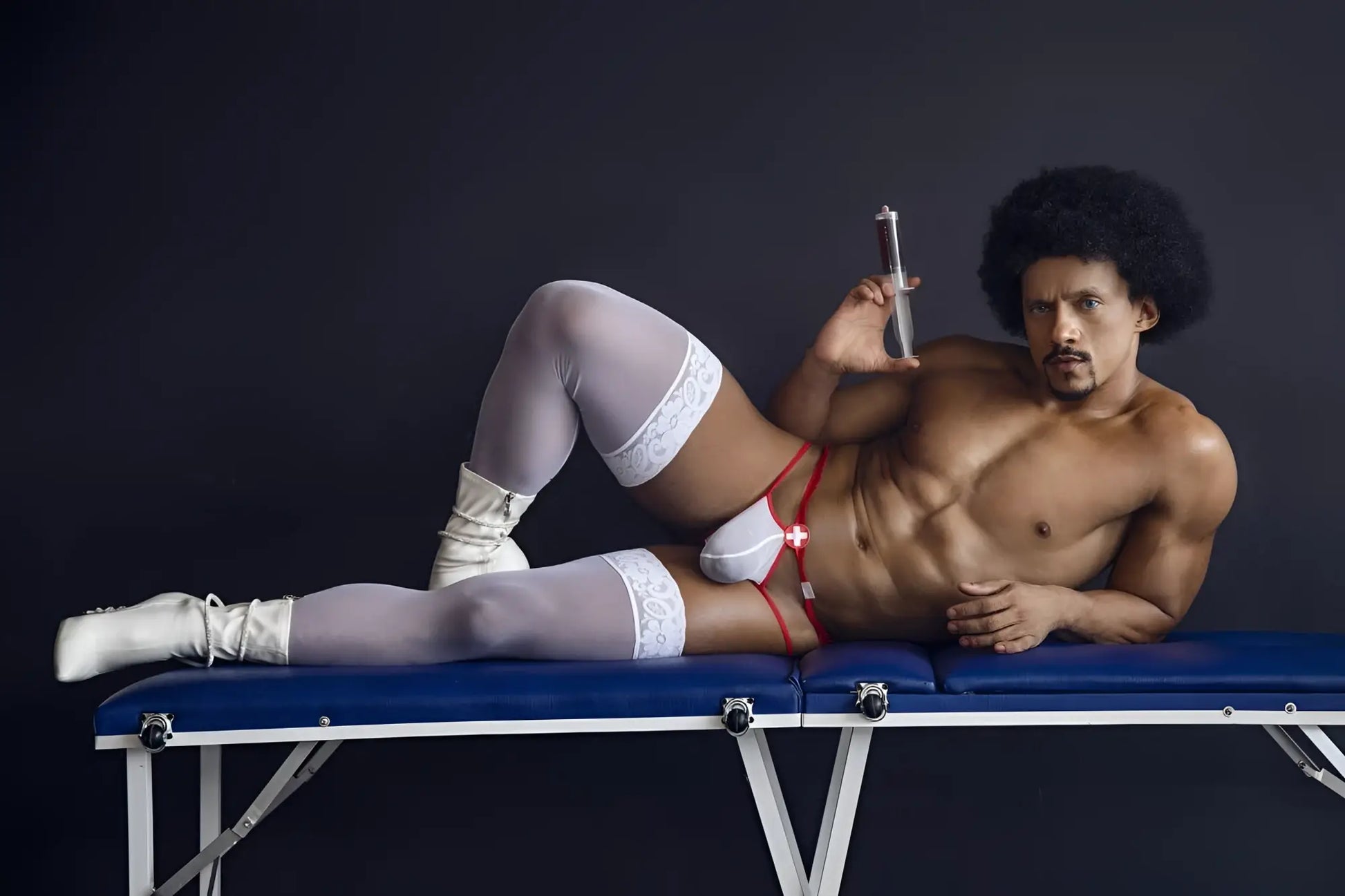 A muscular man wearing CandyMan 99833 murse thongs in red and white with white lace thigh-high stockings on a blue table
