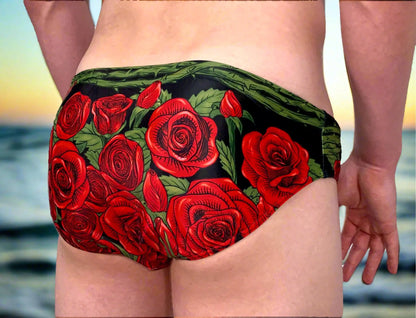 Men's swim briefs in black with vibrant red rose print featuring a snug fit shown by a model at the beach by Maris Equi