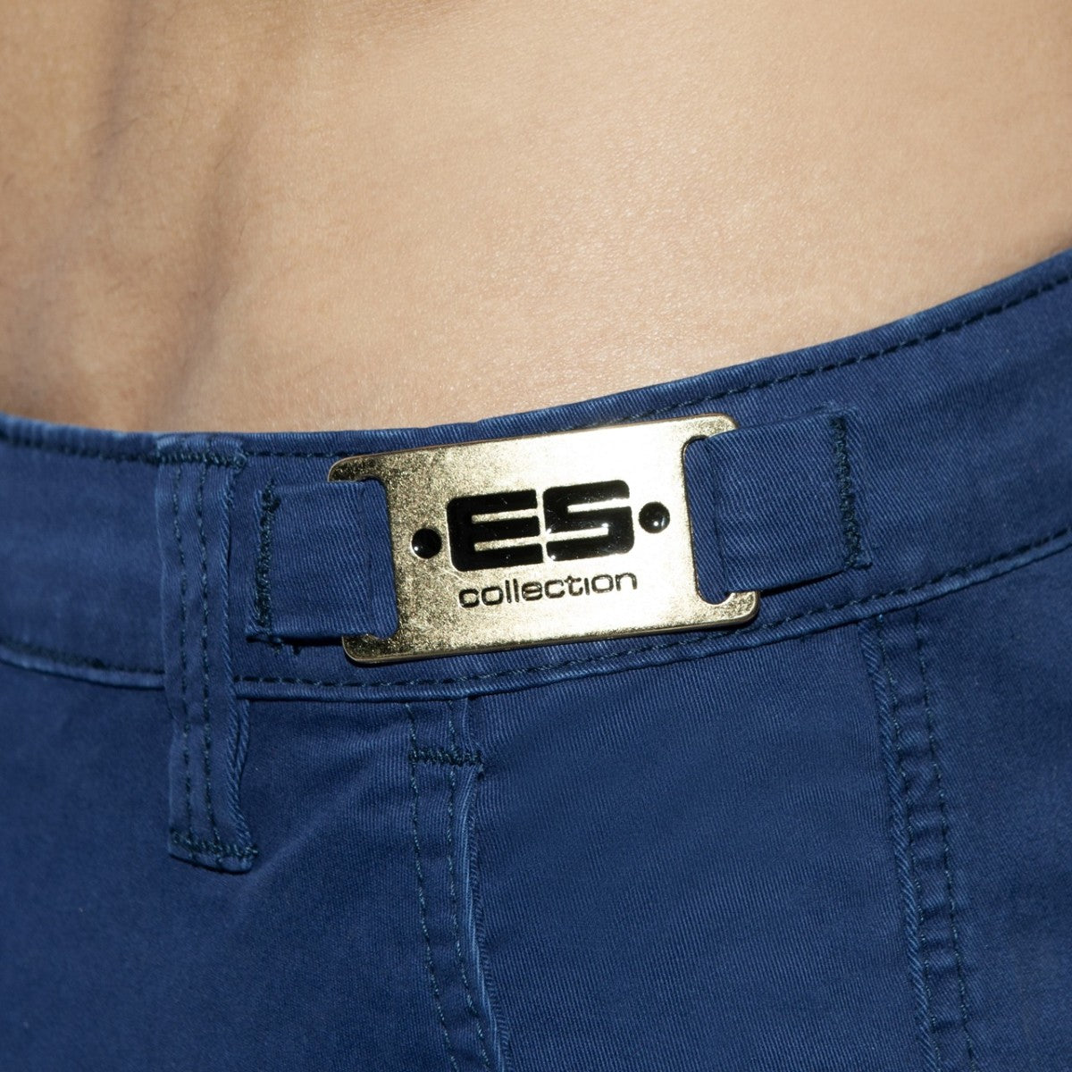 Close-up of blue cargo shorts featuring a gold-toned metal brand plaque on the waistband.
