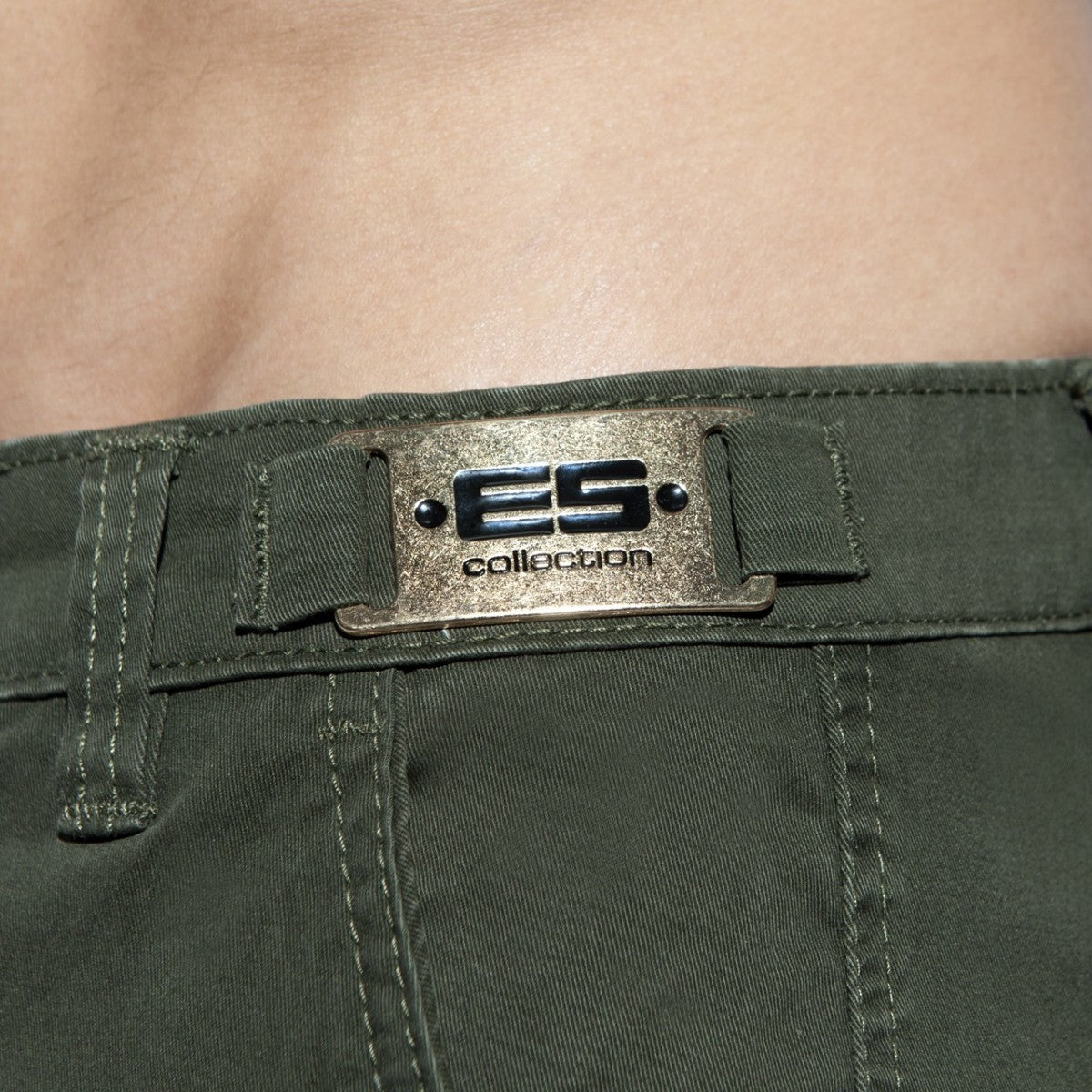 Close-up of olive green cargo shorts featuring a metallic ES Collection brand plate on the waistband.